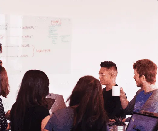 Every single team-member will feel right at home Two men, one with red hair and the other one with short black hair, and two women with long dark hair sitting around a table on a meeting with a board behind them with ideas from a brainstorming on it