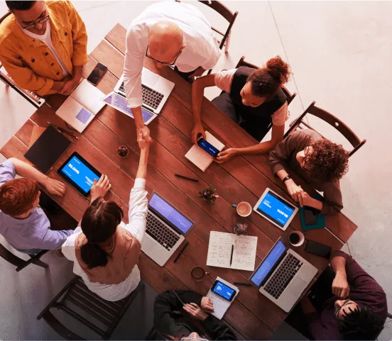Photograph on an unward angle of a rectangular table with 6 people around working on their laptops and taking notes of a meeting