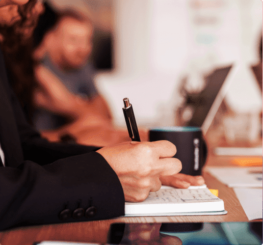 Picture of a woman's hand holding a pen writing on a notebook with a blured meeting on the background