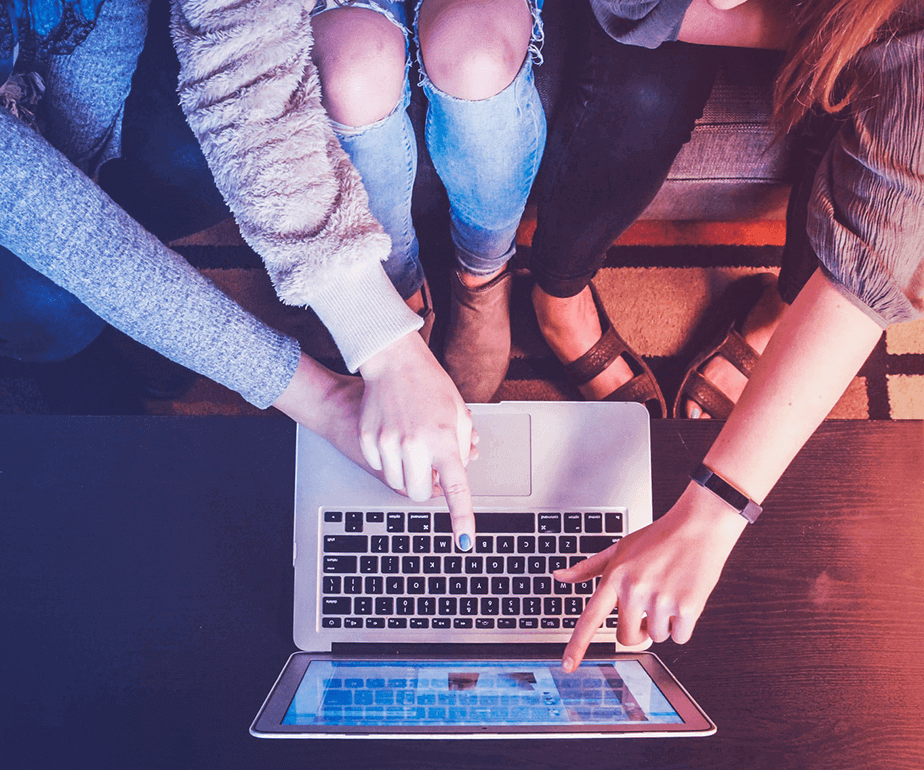 Photograph on an unward angle of 4 women sitting on a couch and pointing at a laptop's screen